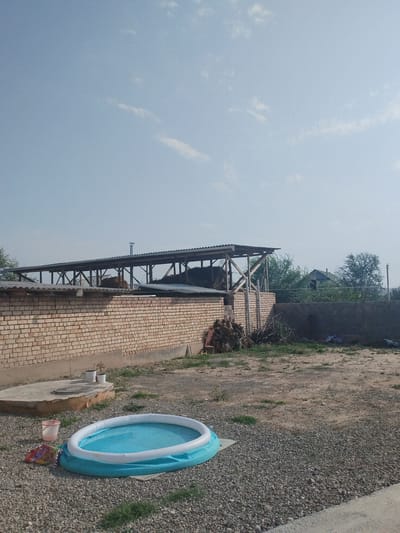 The image depicts a rural or semi-rural outdoor scene, likely in Mady, Kyrgyzstan, under a pale blue sky with sparse, wispy clouds. In the foreground, a small, inflatable blue and white swimming pool filled with water sits on a gravelly surface. To the left of the pool is a red bucket and a multicolored bag. Behind the pool, a low, tan brick wall extends horizontally across the frame. Beyond the wall, a covered, open-sided structure with a corrugated metal roof supported by wooden beams is visible, appearing to store hay or similar materials. Further in the background, trees and the roof of a house can be seen. The ground is a mixture of gravel, dirt, and sparse vegetation. The overall impression is one of a simple, functional outdoor area, possibly a yard or farm.