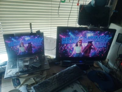The image depicts an indoor scene within the city of Lebedyn, Ukraine, featuring two active monitors on a cluttered desk, positioned in front of a window covered by horizontal blinds.

**Screens and Content:**
Two electronic screens display identical content. The screen on the left is part of a disassembled laptop, its base covered with various items. The screen on the right is a standalone monitor. Both screens show a vibrant, neon-lit image or video frame with the text "НЕ МАРАКУМБА MIX" (NE MARAKUMBA MIX). The imagery includes a disco ball and confetti. Two individuals are featured:
1.  A female with light skin, long brown hair, wearing a white crop top, blue jeans, sunglasses, and white bunny ears, smiling slightly while looking forward.
2.  A male with darker skin, dark hair, wearing a dark top and a dark cap, looking forward with a serious expression.

**Desk Surface (Left):**
The base of the laptop is covered by a transparent plastic sheet, beneath which are various small objects. These include what appear to be electronic components, screws, wires, and at least two compact discs or DVDs. A black, rectangular electronic device, possibly an external drive, is also visible. Below the laptop, a white ceramic cup or bowl rests on a white surface, and a small notebook with visible writing is present.

**Desk Surface (Right):**
The area in front of the standalone monitor is also cluttered. A dark-colored keyboard, possibly wrapped in clear plastic or tape, is angled across the desk. A blue computer mouse is situated to the far right. A small, black rectangular object, possibly a speaker or webcam, is directly in front of the monitor. Various papers and cables are scattered across the surface. A device on the far right displays a red indicator light.

**Background and Environment:**
A window in the background is covered by white horizontal blinds, allowing diffused natural light into the room. Above the monitors, multiple cables and wires hang, some secured with green and white tape. A dark, rectangular object, possibly another monitor or speaker, is visible mounted or placed above the right monitor. The overall environment appears to be a personal workspace or room, characterized by a concentration of electronic equipment and general disarray.