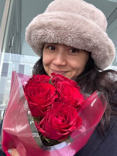 A female subject is depicted from the chest up, centered in the frame, holding a bouquet of red roses. She has dark hair, a medium skin tone, and is looking directly at the camera with a slight smile. She wears a light brown, possibly faux fur, hat that covers her head and ears, and a dark-colored garment. Her right hand, partially visible in the lower left, holds the bouquet. The bouquet comprises at least five fully bloomed red roses, wrapped in a combination of red opaque material and clear plastic film. The background is out of focus, revealing blurred architectural elements consistent with a modern building, including what appears to be glass panels and vertical metallic framing. The scene is located in Sankt Anton am Arlberg, Austria.