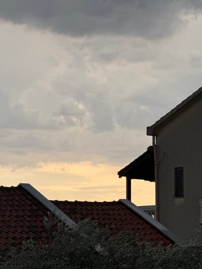 The image depicts a rooftop view of buildings against a cloudy sky. The perspective is from a slightly elevated position, looking across rooftops. In the foreground, a dark tiled roof is visible, with a portion of an olive tree's foliage on the right. Behind this, another building's grey roof extends into the frame, and a small dark-shuttered window is visible on its upper section. A triangular dark awning or roof extension juts out from the side of the second building. The sky is filled with a mix of dark, overcast clouds on the left and lighter, yellowish clouds that suggest sunset on the right. The overall impression is one of approaching dusk, with diffused light. There is no visible text. The scene is set in Trap, Montenegro.