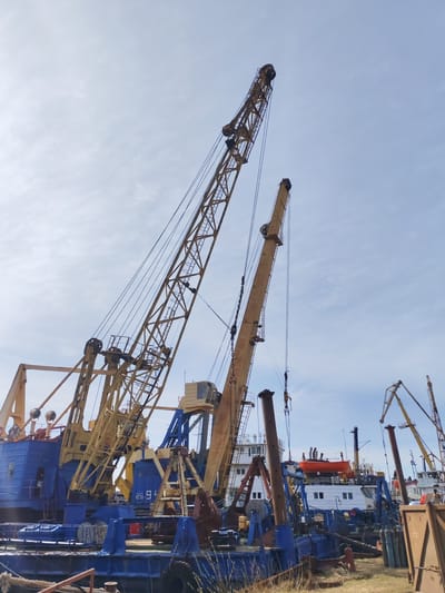 The image shows a harbor scene in Rybinsk, Russia. There are several large, stationary cranes on a blue barge or dock. These cranes are yellow, and their booms are angled upward towards the partly cloudy sky. Other vessels are visible in the background, including a white ship with an orange lifeboat on top. In the lower foreground, there is some vegetation and possibly some discarded construction material.
