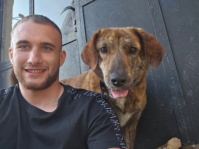A selfie shows a young man with short brown hair, a trimmed beard, and blue eyes. He is smiling at the camera. He is wearing a black t-shirt with white lettering along the sleeves. Beside him is a medium-sized brown brindle dog with floppy ears and a black collar. The dog is panting with its tongue sticking out. They are positioned in front of a dark grey metal door with visible rivets and a handle. The presence of firewood suggests an outdoor area, possibly Lesidren, Bulgaria.