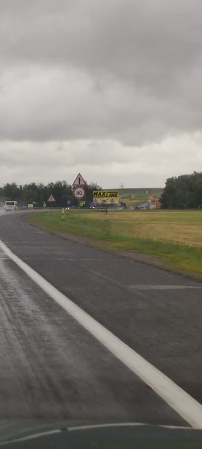 This is a vertical, eye-level, medium shot capturing a highway scene under a heavily overcast sky. The road ahead curves to the left, with a white lane divider clearly visible and wet from rain. The wet asphalt suggests recent or ongoing precipitation.
On the right side of the road, a yellow billboard with the text "MAXLINE" is prominently displayed. The billboard features graphics of what appear to be cartoonish animal figures. Adjacent to the billboard, a small, orange building with windows is visible.
Several road signs are scattered along the roadside. One triangular sign shows a T-junction, while a circular sign beneath it indicates a speed limit of 80. Other signs, some with white and yellow reflective poles, are also present.
In the distance, a line of trees forms the horizon, with some lighter green fields visible between them and the road. A white van is seen driving on the highway to the left, its headlights illuminated. The overall atmosphere is somber and muted due to the cloudy weather, indicating a daytime scene. The location is identified as Vercialishkaŭski Sel'ski Savet, Belarus.