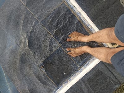 The image captures an overhead view of a person's bare lower legs and feet partially submerged in shallow, sunlit water on a tiled surface in Palasari, Indonesia. The individual's legs are visibly hairy, extending into the water, with their bare feet resting on dark gray, square-patterned tiles. Dark-colored shorts are visible on the upper thighs. The water's surface exhibits distinct wavy patterns and light refractions caused by overhead illumination. A border of white and small blue mosaic tiles runs along the immediate edge of the water, separating it from an adjacent, darker tiled area. A small, dark insect and a tiny piece of white debris are present on the submerged tiles.