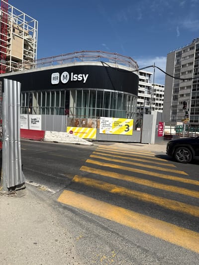 The image depicts a construction site in Issy-les-Moulineaux, France, specifically related to the Grand Paris Express project. A modern building with a black facade and prominent "RER M Issy" signage is visible, suggesting it is a transportation hub or an adjacent facility. The site is secured with metal fencing and scaffolding, indicating ongoing construction work. A large crane, painted red, is present, further emphasizing the active development. Yellow and white striped pedestrian crossing lines are painted on the asphalt road in the foreground. A black SUV is parked on the right side of the image. The sky is clear and blue, and several high-rise apartment buildings are visible in the background. Signs on the construction fencing provide information about the project, including "Grand Paris Express," "gare Issy RER," and "accès pompiers" (firefighter access).