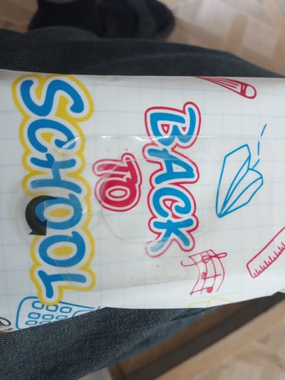 The image shows a close-up of a product packaging with the words "SCHOOL TO BACK" written in large, colorful letters. The text is in a playful, cartoonish font with yellow and blue colors, outlined in red. The background of the packaging is a white grid pattern reminiscent of notebook paper. Scattered around the text are various school-related doodles, including a pencil, a paper airplane, a ruler, and musical notes. A dark fabric, likely clothing, is visible in the foreground and background, obscuring parts of the packaging and suggesting the product is being held or is on a surface. The lighting appears to be indoor, with no strong shadows or indications of time of day. The overall impression is of a product designed for students or the back-to-school season. There is no discernible emotion expressed, and the scene is static. Given the location context of Istaravshan, Tajikistan, the product could be a stationery item, a school supply organizer, or packaging for a school-related snack or treat.