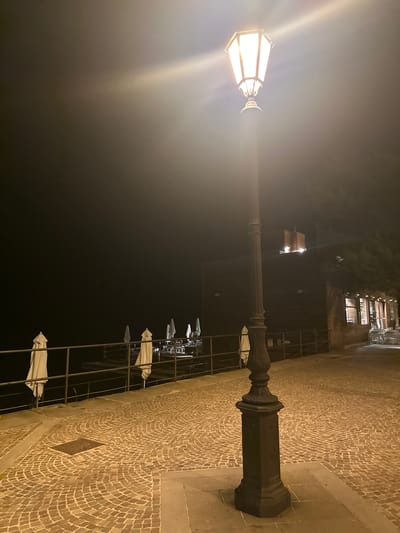 The image depicts a street lamp illuminated at night on a cobblestone walkway. The walkway is next to a metal railing, behind which is a dark body of water. Further back, a building with illuminated windows is visible, along with several closed white umbrellas and tables set up outside. The scene appears to be a waterfront area in Trieste, Italy, possibly near a restaurant or cafe.
