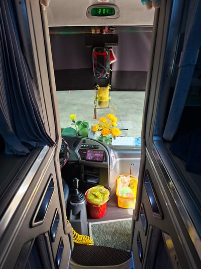This is an interior view of a bus or a similar vehicle, likely taken in Tân Phú 2, Vietnam, looking towards the front cab. The viewpoint is from the aisle, with dark grey or black molded paneling on either side. On the left, a pleated blue-grey curtain is drawn. The front of the bus features a dashboard with a digital clock displaying "2:01", various buttons and switches, a touchscreen display showing a map, and a steering wheel adorned with red and black beads.
Objects on the dashboard include a green hat, a green ornamental gourd, and a vase with artificial yellow flowers. To the right of the steering wheel, there's a red basket filled with yellow plastic bags, and a smaller yellow plastic basket containing some items. A coiled yellow cord is visible near the gear shift. The floor is covered with textured mats. The overall impression is of a well-maintained and somewhat personalized interior, with a mix of functional elements and decorative items. The background appears to be a concrete floor, possibly a garage or parking area.