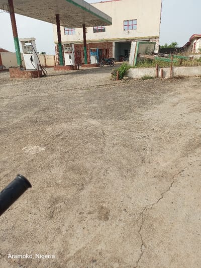 The image shows a deserted gas station in Ado Ekiti, Nigeria, located at coordinates 7.639 latitude and 5.202 longitude. There are three fuel pumps in the foreground, all empty. There are no people visible, but a motorcycle is parked in the background. The gas station is in a state of disrepair with cracked concrete and overgrown vegetation. The building is made of concrete and has a few windows, one of which is broken. The roof is partially covered in metal sheets and has several signs and advertisements. The environment is dusty and dry, with a clear sky overhead. There is a black motorcycle handlebar in the lower left corner of the image, suggesting that the photographer is on a motorcycle.