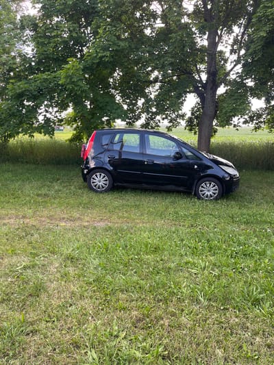 A black hatchback car is parked on a grassy area under a large tree. The car is positioned facing right, with its rear to the left. The area appears to be a rural or park-like setting, indicated by the presence of grass, trees, and a field in the background. The sky is partially visible through the tree canopy, suggesting it is daytime. The car has its headlights on. There are no people visible in the image.