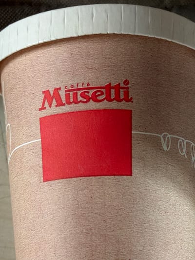 The image is a close-up of a paper cup with the "Musetti caffè" logo printed in red. A solid red rectangle is also printed on the cup. The cup has a white, ribbed rim. The background is dark and out of focus, suggesting it is likely a disposable coffee cup intended for on-the-go consumption, as might be found in various establishments in Piacenza, Italy.