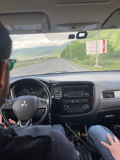 The image shows the interior of a car, looking forward from the passenger seat. A person is driving, with their hands on the steering wheel. The dashboard has a digital display showing the time as 12:38 and a "TIRE PRESSURE MONITOR SERVICE REQUIRED" message. The car is on a road in a mountainous area with a town visible in the distance. A sign by the side of the road reads "LOGISTICS COMPLEX 'MURPHY KAZ' LOGISTIKES COMPLEX 'MEPPHM KAZI'." This suggests the location is in Arsha, Georgia, as Arsha is known for its logistics complex and its scenic mountain setting. The sky is overcast, and the overall impression is of a journey through a scenic, rural landscape.