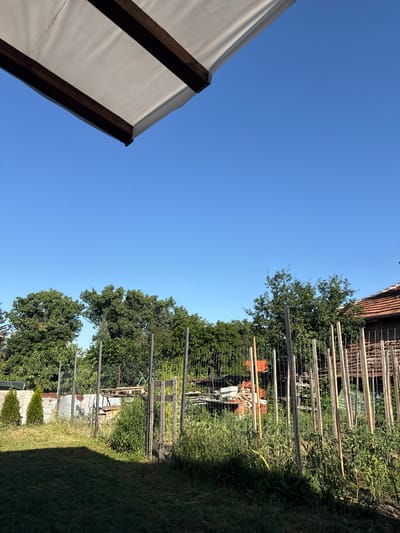 The image shows a backyard scene under a clear blue sky. In the foreground, a portion of a white awning with dark wooden beams is visible at the top left of the frame. The main area of the image depicts a garden with green grass in the lower left quadrant. A fence made of wire mesh and wooden posts runs across the middle ground, separating the lawn from the garden. The garden itself contains various plants, including what appear to be tomato plants supported by wooden stakes. Behind the garden fence, there are stacks of building materials, including bricks and wooden planks. Further in the background, dense green trees form a natural boundary. To the right, a portion of a building with a terracotta tiled roof is visible. Two small conical evergreen trees stand to the left of the fence. The scene is set in the city of Yana, Bulgaria, suggesting a domestic or rural setting. There are no people or animals present in the image.