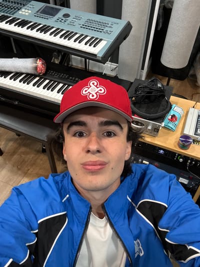 A young man with brown curly hair, wearing a red baseball cap with a white logo, a blue and black jacket, and a white t-shirt, is taking a selfie in a recording studio. He has fair skin and is looking at the camera with his mouth slightly open. Behind him, there are two keyboards, one light blue and one black, stacked on top of each other. To the right of the keyboards, there is a rack of audio equipment, including headphones, a silver device with knobs and buttons, and a small blue and purple cylindrical object. The studio appears to be dimly lit. There is no identifiable context to place this image in the city of Culross, United Kingdom.