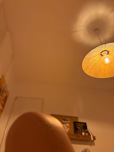 An interior, close-up, low-angle shot focuses on a person's knee and a portion of their thigh in the foreground, with the background depicting a wall and ceiling. A large, round, woven lampshade with a visible lightbulb hangs from the ceiling, casting a circular shadow. On the wall, a white rectangle, possibly a door or panel, is visible. To the left, a framed artwork leans against the wall. Further to the right, a corkboard with two small framed photos and a piece of white lace-like decor is mounted on a wooden shelf, which also displays a framed artwork of a teddy bear sitting in a garden with a small white pot and miniature houses. The overall lighting is warm and yellow, originating from the hanging lamp, creating a cozy ambiance. The scene appears to be in a residential setting, potentially a bedroom or living area. There is no indication of the location being in Benissa, Spain.
