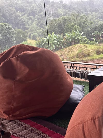 The scene is an elevated outdoor or semi-covered seating area in Ban Paem Bok, Thailand, experiencing heavy rainfall. In the foreground, a large, rust-colored fabric beanbag chair occupies the left, resting upon a patterned mat featuring red, brown, and cream colors, which is laid over artificial green turf. A dark gray square cushion is partially visible beneath the beanbag. To the right, a dark wooden table and a segment of a rope-woven outdoor seating element are present. A black metal railing with horizontal bars defines the edge of the immediate platform. Beyond the railing, a dense tropical forest covers undulating hills, with visible rain streaks indicating a heavy downpour. The vegetation includes numerous banana plants on a lower slope and a variety of green broadleaf trees, interspersed with a distinct light-colored, leafless tree in the mid-ground. A thin, dark vertical support pole extends from the upper center of the frame. No individuals are explicitly depicted.