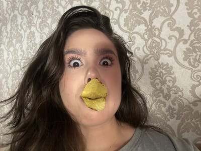 A young woman with dark, wavy hair is pictured in a close-up, self-taken photograph. She has her eyes wide open, looking directly at the camera, and is wearing a significant amount of eye makeup. A potato chip is placed over her lips, giving the appearance of a unique mouth. The background is a patterned wallpaper. The image is taken indoors, with no identifiable landmarks from Raivka, Ukraine.