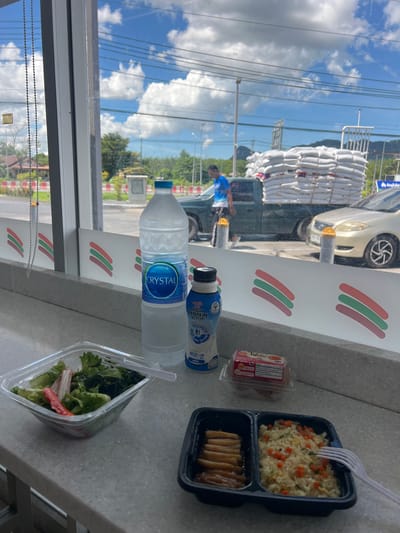 The image shows a close-up of a convenience store counter next to a window. On the counter are several food items and drinks: a black plastic container with rice and carrots, a smaller black container with what appears to be fried chicken strips, a clear plastic container with a salad, a bottle of Crystal water, and a bottle of a blue liquid. A plastic fork is stuck in the rice. In the background, through the window, a busy street scene is visible, with a car, stacks of white bags on a truck, and a person wearing a blue shirt. The reflection in the window shows a bright sky with clouds, suggesting it is daytime. The convenience store's branding, featuring colored stripes, is visible on the counter. A small, rectangular package with text and images is also on the counter to the left of the rice. The overall impression is a quick meal or snack purchased at a convenience store.