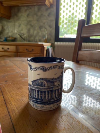 The image captures a ceramic mug lying on its side on a wooden table, illuminated by strong sunlight. The mug is cream-colored with a dark blue interior and features a detailed blue illustration of a classical building, possibly an architectural landmark. Along the side of the mug, the text "SAINT PETERSBURG" is visible, indicating its souvenir origin.
The wooden table surface displays a prominent grain texture and numerous scratch marks, suggesting age and use. Bright, irregular patterns of light, possibly sunlight filtered through a window or leaves, are cast across the table and the mug, creating a dynamic interplay of light and shadow.
In the background, a large window is visible, covered by a light-colored, patterned mesh or curtain with a repeating green, leaf-like motif. Beyond the window, blurred greenery indicates an outdoor setting with a sunny day. To the left and behind the mug, parts of a wooden chair or shelving unit can be seen. Further in the background, a blurry section reveals what appears to be a tiled wall, possibly in a kitchen or utility area, and segments of wooden cabinets or furniture, one with a dark handle. The overall scene suggests a domestic interior, likely a kitchen or dining area, in Courpière, France, during daylight hours. No specific activity or people are depicted; the image presents a still life.