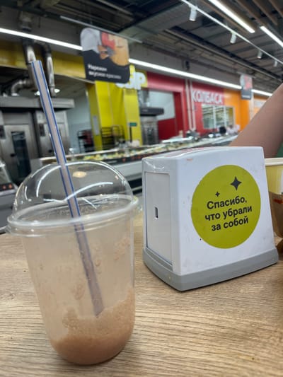 The image depicts a close-up view of a table in what appears to be a cafe or food service area within a supermarket in Volzhsky, Russia. In the foreground, a partially consumed beverage in a clear plastic cup with a lid and straw sits on a wooden table. To the right of the cup is a white napkin dispenser with a green circular sticker. The sticker has a black star-like symbol at the top and Cyrillic text that reads "Спасибо, что убрали за собой" (Thank you for cleaning up after yourself). The background reveals the interior of a commercial establishment, possibly a supermarket aisle, with bright lighting, shelving units, and signage. Notably, a sign with "КОЛБАСА" (Sausage) is visible in the background, suggesting a food retail environment. The overall scene is a casual snapshot of a personal moment, likely taken by a customer.