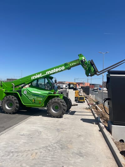 In Alzey, Germany, a bright green telehandler is parked on a paved surface. The telehandler's boom is extended upwards and slightly to the right, with its forks positioned near a tall, black electrical charging station. The charging station is mounted on a concrete base and has a thick black cable extending from it. The telehandler has "mi...mateco" written on its boom and "mateco" on its side, along with the model number "TS 1072 4x4 31101030" and the weight capacity "7,2 to.". Two large, knobby tires are visible on the left side of the telehandler. In the background, several trucks, construction equipment, and buildings are visible under a clear blue sky with a faint contrail. The ground is a mix of concrete and dirt, with some construction debris scattered around.