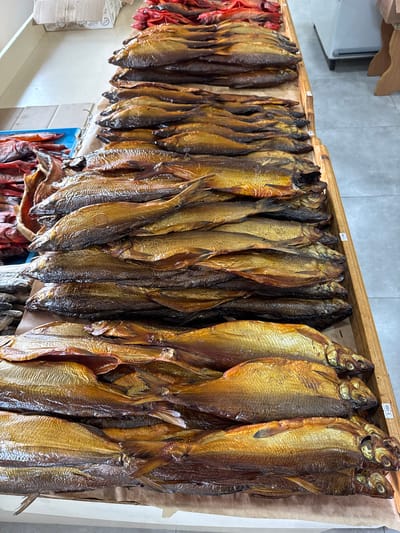 The image shows a display of smoked fish at a market, likely in Tsovagyugh, Armenia, based on the prompt. The fish, predominantly golden-brown, are hanging in a row, attached to a wooden frame. The fish appear to be densely packed together. Some smaller red fish are visible on the left of the photo, but the main focus is on the larger golden-brown fish. The background shows a plain wall. A price tag can be seen on the wooden frame.