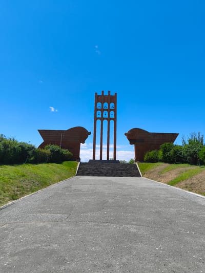 The image shows a monument against a clear blue sky in the city of Araks, Armenia. The monument features a tall, three-arched structure with bells, flanked by two large, abstract, wing-like sculptures made of a reddish-brown material. A wide, paved pathway leads up to a set of concrete stairs that ascend to the monument. Lush green grass covers the slopes on either side of the pathway and stairs, with some trees and bushes visible in the background. The overall scene is peaceful and grand, evoking a sense of historical significance.
