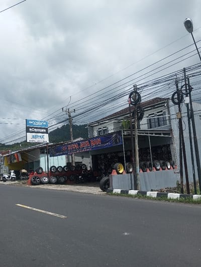 A tire shop named "Syifa Jaya Ban" displays various tires and rims along the front of its building. A banner with the store's name hangs above the entrance. Multiple power lines run across the sky. The shop is located along a paved road with a yellow line. Across the street, a utility pole and street light stand tall. The sky is overcast with a backdrop of distant mountains.
