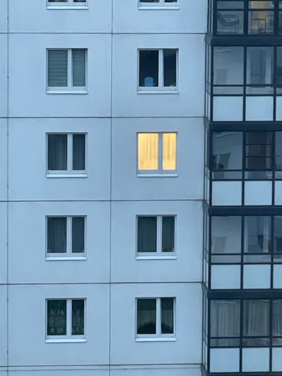 The image displays a section of a multi-story building in the city of Novogorelovo, Russia. The building features a facade with rows of windows. On the left side, there are several windows with light blue or gray curtains. One window in the middle row on the left is illuminated with a warm, yellow light, suggesting interior lighting. To the right of these windows, there is a section of the building with a glass-enclosed balcony or a series of windows that form a glazed facade, divided by dark frames. The sky appears overcast, contributing to the muted lighting of the scene.