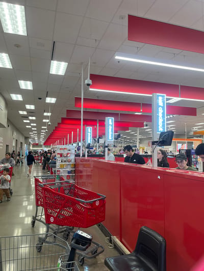 The image displays the brightly lit self-checkout area of a Target retail store in El Paso, United States. Multiple self-checkout stations are arranged in a long row, defined by red barrier panels and illuminated by blue "Self checkout" signs suspended overhead.
In the foreground right, two individuals are actively using self-checkout kiosks. A male, wearing a dark t-shirt and sunglasses on his head, operates one kiosk, while a female with glasses is visible at an adjacent station. Further into the midground, another male in a light-colored top is at a self-checkout kiosk.
On the left side of the scene, a woman pushes a red Target shopping cart, accompanied by a young child walking beside her. Further down the aisle, additional customers are visible walking or queuing. Two empty red Target shopping carts are positioned prominently in the mid-foreground, facing left. A smaller, gray cart is partially visible in the immediate foreground. Merchandise displays, including impulse-buy items, are situated near the checkout lanes.
The ceiling consists of a white grid system with numerous recessed fluorescent light panels and linear LED strip lights, providing bright illumination. Multiple white dome security cameras are mounted on the ceiling, overseeing the area. Red decorative banners are suspended across the ceiling, characteristic of the Target brand. The flooring is a light-colored, polished surface. A black chair, likely for staff, is partially visible behind the red barrier in the bottom right corner.