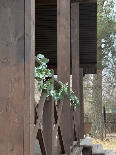 This image captures a section of a dark brown stained wooden structure, identified as being in Dubki, Russia. The structure features vertical support beams and a decorative railing with an X-pattern design. Faux eucalyptus leaves and small white flowers are draped along the railing. Overhead, a dark, horizontally grooved soffit is visible. To the lower right, two wooden benches are integrated into the structure. Beyond the structure, the scene transitions to an outdoor environment dominated by numerous deciduous trees with bare branches, suggesting a dormant season. A large tree with textured bark stands prominently in the middle ground. The ground is covered with dry, light-colored vegetation. In the far background, a chain-link fence or wire mesh structure is discernible, behind which a blue and yellow element, possibly part of a sports court or recreational equipment, can be seen. No individuals are present in the frame.
