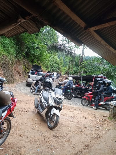 The image shows a dirt road lined with parked vehicles and motorcycles.  It is taken from under a corrugated metal awning. The location is a rural area near Cisewu, Indonesia, at coordinates -7.411 latitude and 107.542 longitude.  The vehicles are a line of motorcycles and two cars, one of which is a white SUV.  The motorcycle closest to the camera is a white Yamaha scooter.  Other motorbikes are red, black, and blue. The SUV is parked behind the motorcycles.  There are several people in the image, including people on motorbikes, and people standing by vehicles. All are adults, and they are dressed casually. The scene is set against a lush green backdrop of trees and foliage. The ground is muddy and unpaved.  There is a large wooden structure visible in the background, as well as a group of people standing in the distance.  The scene is likely one of a parking area or gathering place.