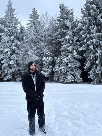 A male subject stands in a snow-covered field in Le Hohwald, France. He is positioned slightly left of center, facing to the right with a neutral expression. He wears a black beanie with a white Nike logo, a black quilted puffer jacket over a light gray shirt, black gloves, and dark pants. His shoes, partially visible under the snow, are dark with red and white accents. The ground is covered in deep, undisturbed white snow, with some foot impressions present. In the background, a dense forest of coniferous and bare deciduous trees, heavily coated in snow and frost, extends across the scene under an overcast, light gray sky.