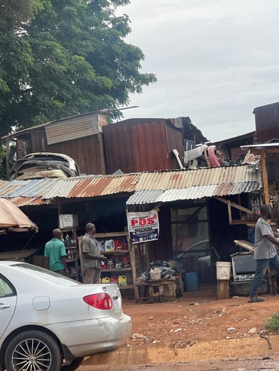 The image depicts a scene in Karshi, Nigeria. The main subject is a small, makeshift roadside shop constructed of corrugated metal and wood. It appears to be an automotive repair or parts shop, as evidenced by car parts and engine oils displayed. Several people are present, including customers and workers. One individual stands on an elevated platform within the shop, while others are visible near a parked white car on the right. A sign for "Moniepoint POS Machine" is prominently displayed, with local phone numbers. Trees are seen in the background. The environment is characterized by a dusty, unpaved street.