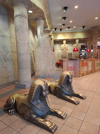 This is a photograph of an indoor Egyptian-themed restaurant in the city of Trafford, United Kingdom. The scene features two large, bronze-colored sphinx statues in the foreground, positioned on a tiled floor. Behind the sphinxes, there are tall, stone-like columns decorated with hieroglyphs. In the background, a wooden counter with signage advertising food and drinks, such as "PIZZA PASTA and more HOT EAT DELICIOUS." There are also statues of human figures and red lamps hanging from the ceiling. The overall ambiance suggests a themed dining establishment.