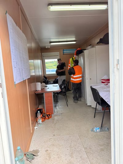The image depicts the interior of a temporary site office, possibly a portable cabin, located in Jodłownik, Poland. The room is functional and appears to be in active use for construction or site management.

In the foreground, on the left, two plastic water bottles are visible, one partially filled. Next to them, a pair of work gloves rests on the floor.

The interior walls are clad in wood-effect paneling. A large technical drawing or blueprint is pinned to the left wall. In the center of the room, a small, orange and grey desk stands with a roll of toilet paper and a pen on its surface. A black office chair is positioned behind it.

Two men are present in the scene. One man, wearing a dark shirt and light-colored pants, stands near a window to the right of the desk, holding what appears to be a mobile phone or small device. He is looking down at it.

Further into the room, a second man, wearing a dark long-sleeved shirt, dark pants, and a bright orange high-visibility vest with reflective stripes, is standing near a row of white lockers. He is holding a dark helmet in his hands.

On the floor near the lockers, a black backpack is visible. Behind the man in the high-visibility vest, the lockers appear to be industrial-style. On top of the lockers, dark fabric items are stored.

Further to the right, another black office chair is positioned at a table which is covered with papers, including what looks like another technical drawing. On a shelf above this table, a stack of red and white items is visible.

A plastic water bottle lies on its side on the floor in the right-hand portion of the image. A portable air conditioning unit is mounted high on the wall towards the back of the room, to the left of the lockers. The floor is a light-colored, speckled material, possibly linoleum or concrete, showing some signs of dirt and wear. The scene is lit by fluorescent strip lights on the ceiling. The entrance to the cabin is visible on the far right, with a white frame. The window in the background offers a view of an outdoor landscape with greenery.
