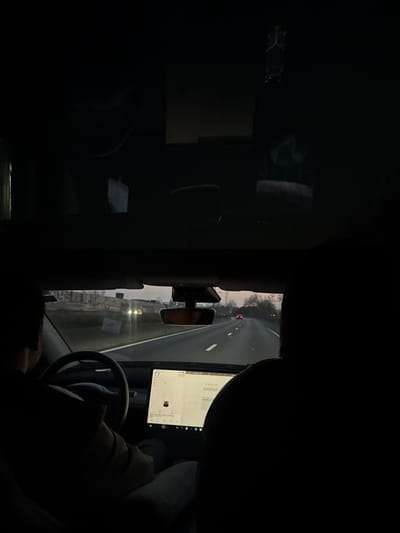 The image captures the interior of a vehicle, consistent with a Tesla, in motion on a multi-lane highway in Rosny-sous-Bois, France, during twilight or early evening. The vantage point is from the rear passenger area, looking forward. A driver is visible in the front left seat, with hands on the steering wheel. The silhouette of a front passenger is discernible on the right. The vehicle's central touchscreen display shows a navigation map on the left and "TESLA CINEMA" on the right, indicating a 77% battery charge. A dashcam is mounted below the rearview mirror. Outside, the highway extends into the distance, marked by lane lines. Other vehicles are present ahead, distinguished by red taillights and distant white headlights. To the left of the road, a concrete barrier with indistinct markings or graffiti is visible, backed by faint structures. To the right, trees line the roadside against the fading light of the sky. The vehicle's panoramic glass roof or upper windshield reflects internal elements.