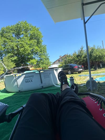 In Suvorovo, Bulgaria, on a sunny day, a person is relaxing in a chair in a yard. The person is wearing black pants and black and white sneakers, with their legs extended. In the background, there is a large above-ground swimming pool with the "Bestway Steel Pro MAX" logo visible on its side. A black car is parked behind the pool, and several trees with green foliage are scattered throughout the yard. A section of a white canopy structure is visible in the upper right corner of the image. The ground is covered with artificial green turf. The sky is clear and blue.