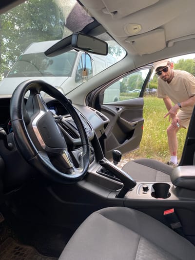The image captures an interior view of a car's cabin, looking towards the driver's seat and out the open passenger door. A man, wearing a light-colored t-shirt and shorts, is visible outside the car, leaning in. He has sunglasses on and is making a gesture with his hand. His right leg has a visible injury with a bandage. The car's interior shows a steering wheel with "AIRBAG" text, a dashboard with a screen, a gear shift, and a center console with cup holders and a control knob. A rear-view mirror with a small air freshener is mounted on the windshield. Outside the car, greenery and a glimpse of a white vehicle are visible, suggesting an outdoor setting. The scene appears to be during the daytime, with natural light filtering into the car. The overall impression is a casual moment, possibly between taking a break or discussing something. The visible text includes "AIRBAG" on the steering wheel. The location is identified as Bielsko-Biala, Poland.