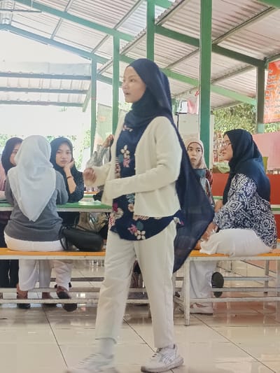 A young woman wearing a dark blue hijab, floral top, and white cardigan walks in front of a group of seated women. Some of the women are wearing hijabs, while others have their hair covered in headscarves. The women are sitting at picnic-style tables in an open-air setting with a metal roof. The woman is wearing white pants and white sneakers.
