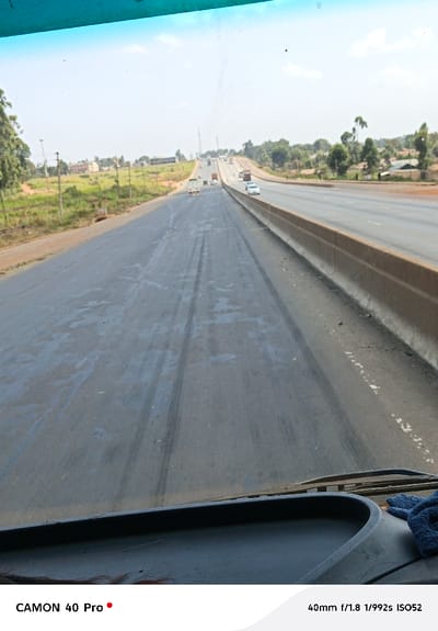 A long, straight road stretches into the distance, marked by tire tracks. A barrier separates the road from another higher road to the right, where cars are driving. The view is captured from inside a vehicle, with part of the dashboard visible. Greenery and buildings line the sides of the road. The sky is bright, suggesting a clear day.