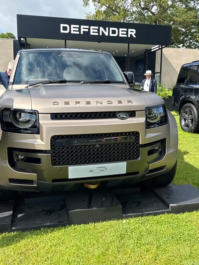 A brown Land Rover Defender is parked on black platform mats on a grassy area. The vehicle is on display under a large black awning with the word "DEFENDER" written in white on it. There is a person in a white hat and a black jacket standing to the right of the vehicle, looking at it. The vehicle is facing the camera, and the front grille and headlights are visible. The Land Rover emblem is centered on the front of the vehicle.  The coordinates provided are for the town of Bramham, West Yorkshire, United Kingdom.  However, the exact location cannot be determined from the image.