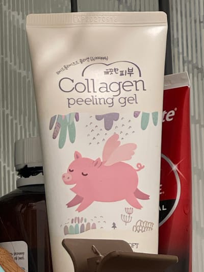 This is a close-up, eye-level shot of a skincare product tube, specifically a "Collagen peeling gel," positioned horizontally. The tube is predominantly white with purple text and a cartoon illustration of a pink pig with wings. To the left of the tube, the text "EXP20270918" is visible, indicating an expiration date. The background consists of a tiled wall, suggesting a bathroom or vanity setting. Above the white tube, a sliver of a red and white toothpaste tube is visible, with the brand name "Colgate" partially obscured. To the right of the white tube, a brown object, possibly a holder or another bottle, is partially visible. The lighting is even, indicating indoor lighting. The overall scene conveys a sense of personal care and hygiene. The visible text is in English and Korean, with the main product name "Collagen peeling gel" clearly legible. The Korean text translates to "Hydrolized Collagen (5,000ppb) for clean skin."