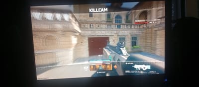 The image shows a screenshot from a video game, likely a first-person shooter, displaying a "KILLCAM" sequence. The environment appears to be an urban setting with stone architecture, reminiscent of European cities, though no specific location in Karviná, Czechia, can be identified from the provided image.

In the foreground, a player's view is dominated by a light-colored submachine gun, the "CARBON 57," with a scope. The in-game interface displays information such as "ATTACKERS: 1", the attacker's username "[0cr]GobeLamoistp" along with their in-game stats (300 kills, 13 deaths). Another player was killed by this attacker, indicated by "KILLED BY: [0cr]GobeLamoistp #9231842". There's an option to "REPORT PLAYER" and "COPY BUILD". The player is at "LEVEL 41".

The background features a building with arched windows and ornate stonework, a large red door, and some greenery. The scene is well-lit, suggesting daytime gameplay. The overall image quality is slightly grainy due to the photograph of a screen. There are no actual people or interactions visible, only in-game representations.