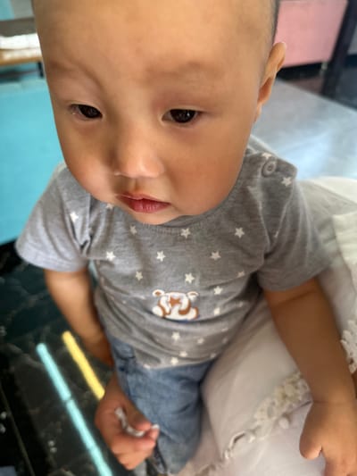 The image shows a close-up of a young child, likely a toddler, looking down. The child has fair skin, dark eyes, and short dark hair. They are wearing a grey t-shirt with white stars and a cartoon bear graphic, along with denim jeans. The child's left hand is visible, holding a small white object. The background is blurred, but hints of blue and pink furniture, as well as a dark, reflective surface with colored lines (possibly a floor or table), can be discerned. The lighting suggests an indoor setting, possibly a home or a child-friendly establishment. The specific location in Ынтымак, Kyrgyzstan, cannot be determined from the visual information alone.