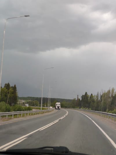 The image shows a road in Chusovskoye Urban Settlement, Russia, under a cloudy sky. A gray asphalt road curves through a landscape with trees on either side. A white truck is visible on the road in the distance. Street lights line the road. The view is likely taken from inside a vehicle, as the windshield is visible at the bottom of the frame.