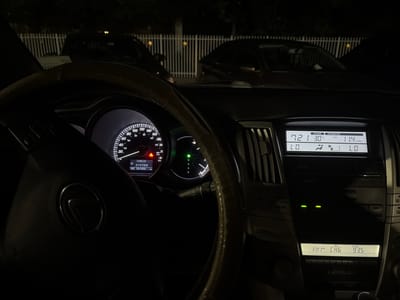 This is a nighttime interior shot of a car, likely in Aventura, USA. The focus is on the dashboard, with the steering wheel prominently in the foreground, displaying the Lexus logo. The instrument cluster shows the speedometer reading 20 mph, with other gauges indicating engine temperature and fuel levels. The car's odometer displays "281456 km".
To the right of the steering wheel, the car's infotainment system is visible. The display shows "OUTSIDE 721 30 C", suggesting an outdoor temperature of 30 degrees Celsius and possibly a time of 7:21. It also shows "INFORMATION AVG 11.4 L/100km", indicating average fuel consumption. Below this, climate control settings are displayed, with icons for temperature and air flow. The radio display shows "FM1 CH6" and "ST MSG 93.5", indicating the car is tuned to FM radio station 93.5.
Two cars are visible through the windshield, parked behind a white picket fence. The overall lighting suggests it is dark outside, consistent with nighttime. The scene appears to be a parked car, with no activity taking place. The image is sharp and well-lit internally, contrasting with the dark exterior.