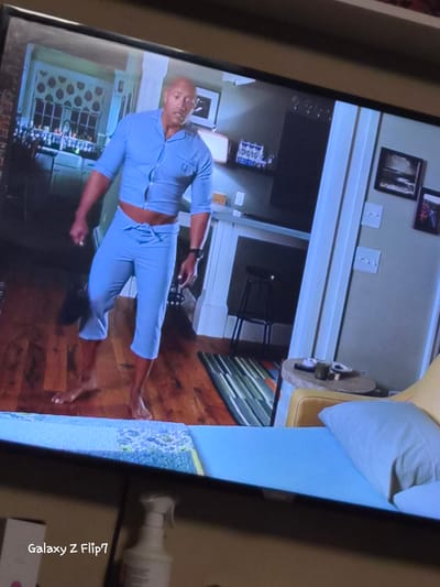 The image displays a photograph of a screen, likely a television or monitor, showing an indoor scene. The screen is tilted diagonally within the frame.

On the screen, the primary subject is a bald man, possibly Dwayne "The Rock" Johnson, standing barefoot on a wooden floor. He is wearing a light blue, two-piece outfit resembling pajamas or a leisure suit, consisting of a short-sleeved top and capri-length pants. The man is looking slightly to his left, with a neutral expression.

The background on the screen depicts a home interior. To the left of the man, there's what appears to be a large window or decorative panel with a patterned, possibly honeycomb-like, design, with shelves or a rack beneath it holding numerous bottles or containers. Further back, beyond the man, various pieces of furniture are visible, including a dark-colored chair and a table. Framed pictures hang on the light-colored walls. To the right of the man, a section of a bed or couch with a light-colored pillow is visible.

Outside the screen's frame, to the right, a white spray bottle is partially visible. At the bottom right corner of the original image, text "Galaxy Z Flip7" is clearly visible, indicating the device used to capture the photo of the screen. The overall lighting suggests an interior setting, possibly during the daytime or with artificial light.