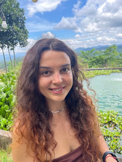 A medium shot captures a young woman with long, curly brown hair, smiling at the camera. She is wearing a brown top and a beaded necklace. The background suggests an outdoor setting, possibly a resort or natural park. Lush green vegetation and a body of turquoise water with lily pads are visible. In the distance, rolling hills are covered in trees, under a partly cloudy blue sky. The overall scene evokes a sense of relaxation and natural beauty, with a hint of an exotic location.