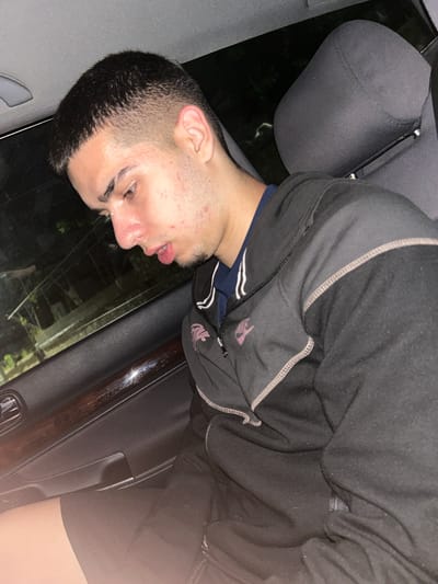 A young man with short dark hair and light skin is positioned in a car. He is wearing a black zip-up hoodie over a blue shirt. He appears to be either sleeping or unconscious, with his mouth slightly open. The car's interior is visible, including a gray headrest, door paneling, and what appears to be a door handle. Outside the car window, there is darkness with some indistinct light sources, possibly suggesting a nighttime setting. The location, Pripek, Bulgaria, is mentioned as context.